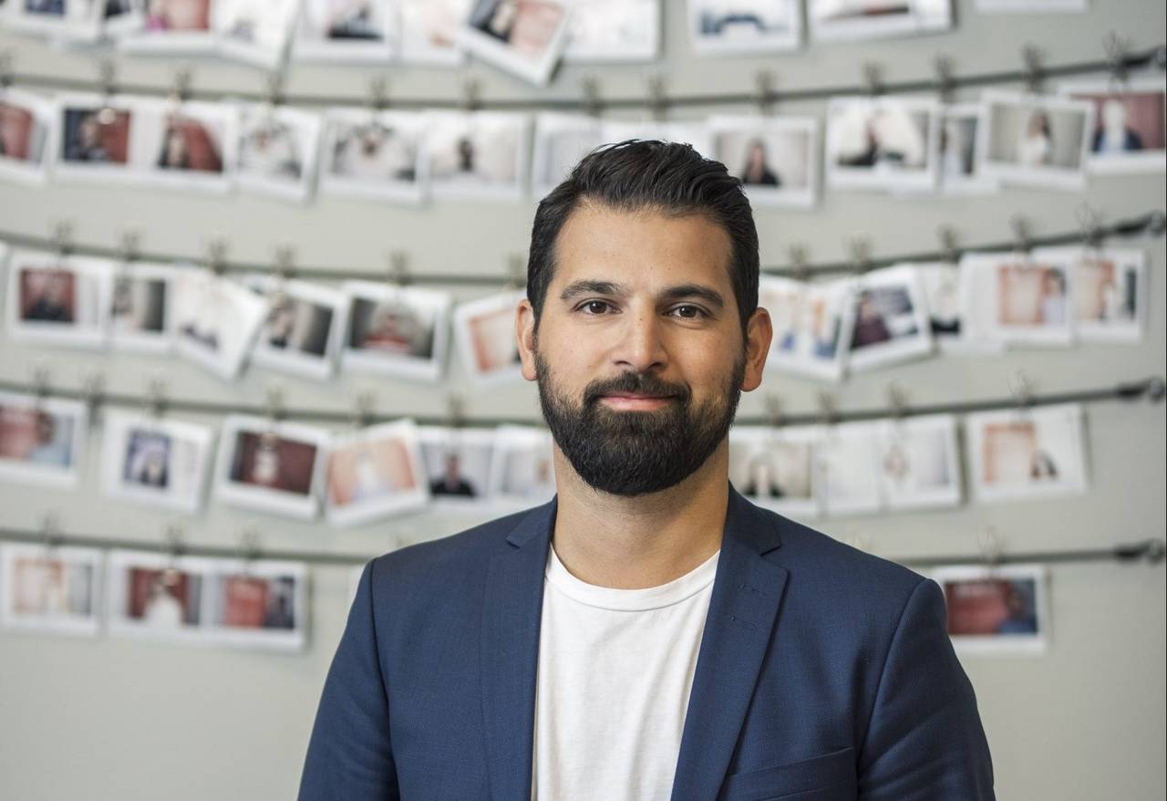 Farzad Golchin bör blå kavaj över vit tröja. Han har mörkt hår och skägg. Bakom honom syns band där mängder av fotografier hängts upp.
